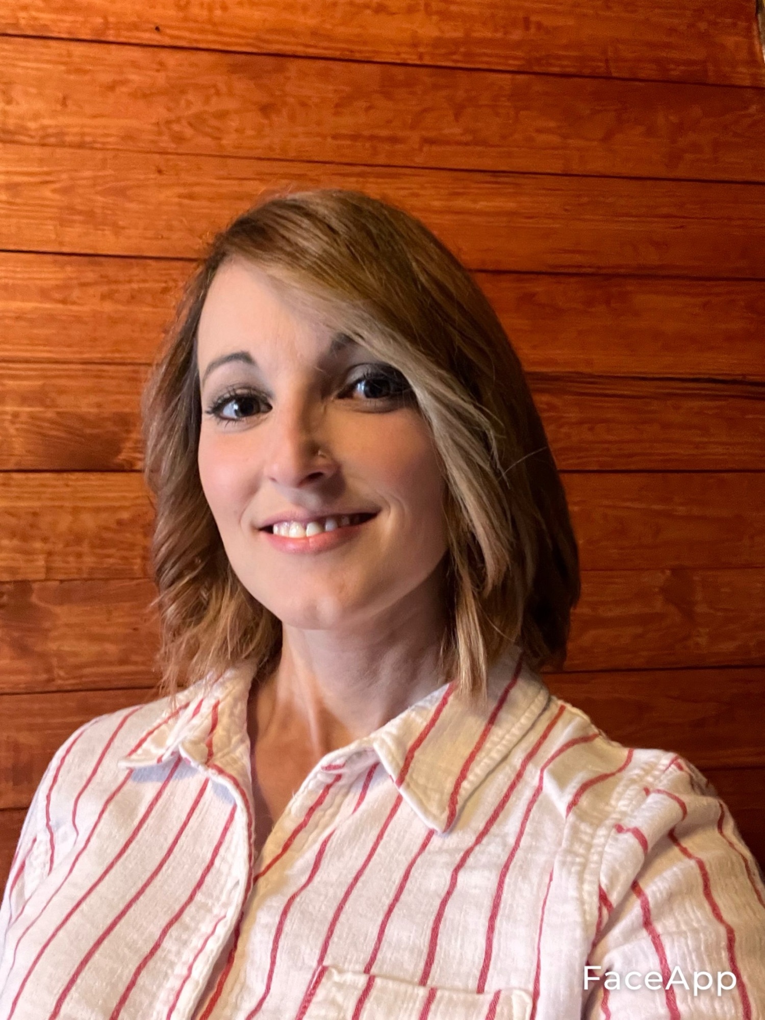 A smiling woman with shoulder-length hair wearing a white shirt with red stripes, standing in front of a wooden wall.